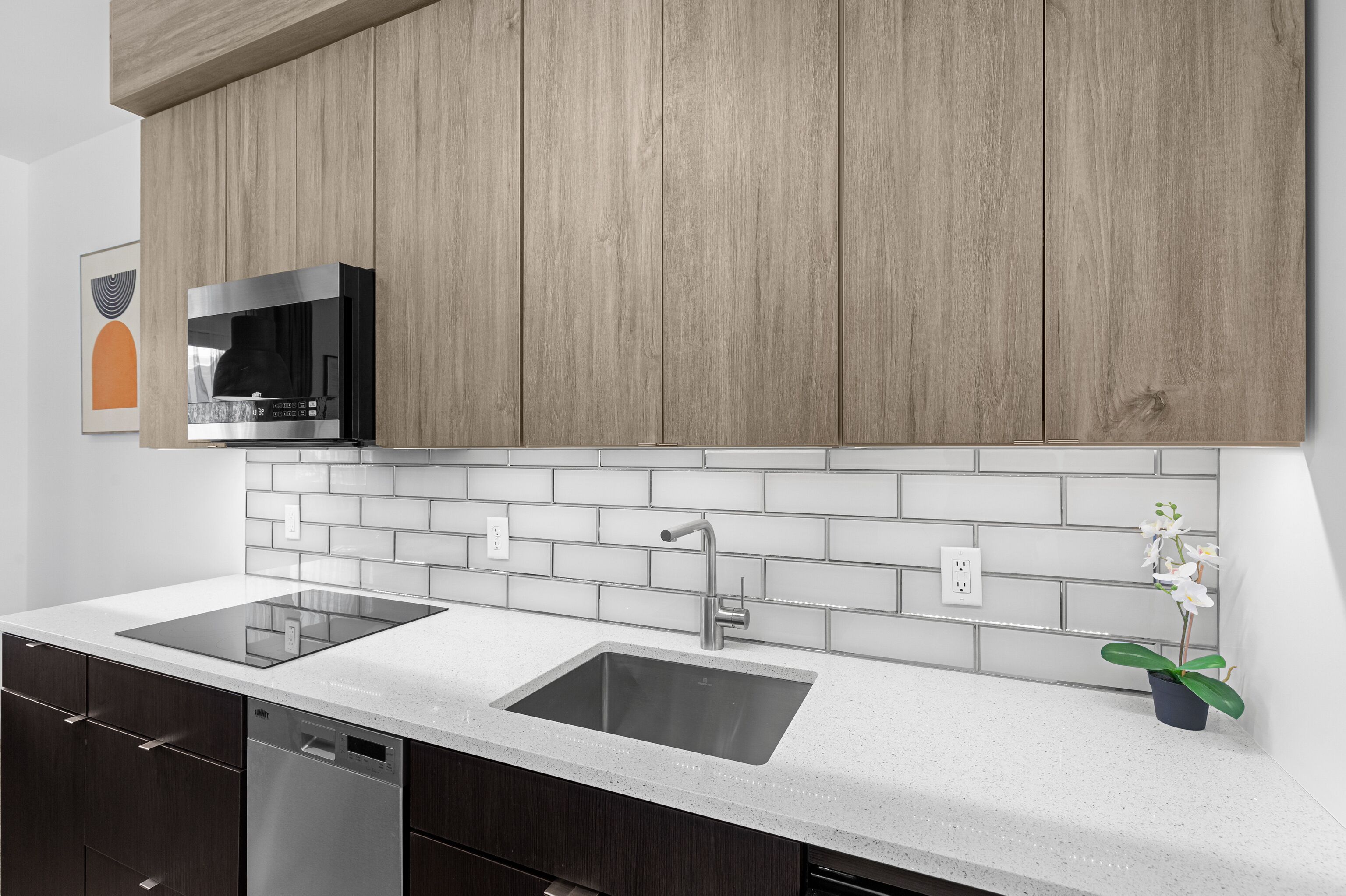 A modern kitchen with wooden cabinets, a stainless steel microwave, a double sink, and a potted orchid.