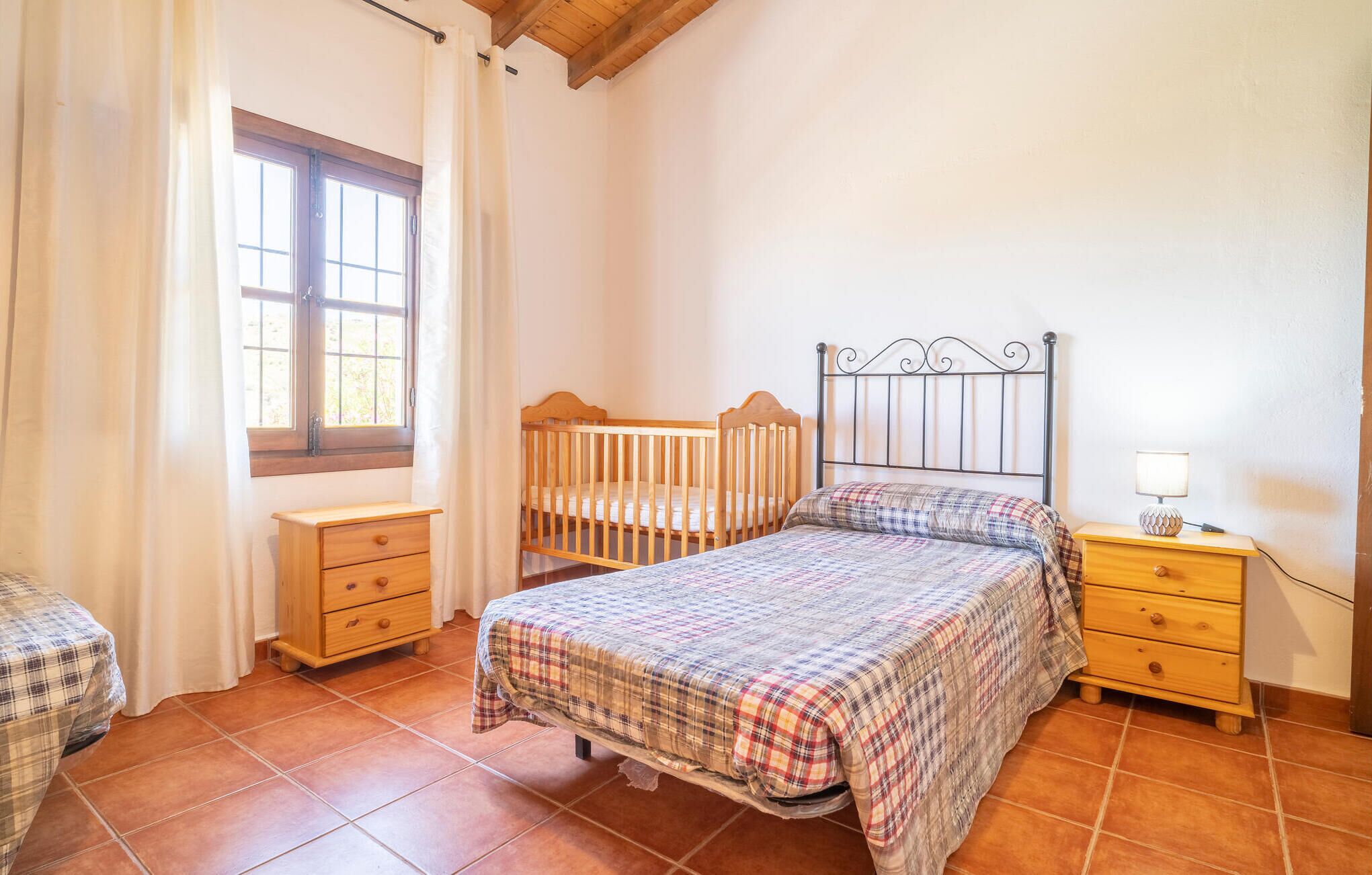 Een slaapkamer met een houten ledikant, een tweepersoonsbed met een geruit sprei, een nachtkastje met een lamp en een raam met gordijnen.