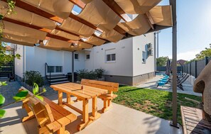 Eine Außenpatio mit Holztischen und -bänken, einem überdachten Pergola und Blick auf ein Haus mit weißer Fassade und einem eingezäunten Garten.