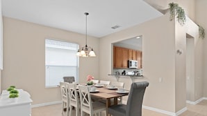 A dining area with a wooden table, white chairs, and a chandelier. A kitchen is visible in the background with wooden cabinets and a microwave.