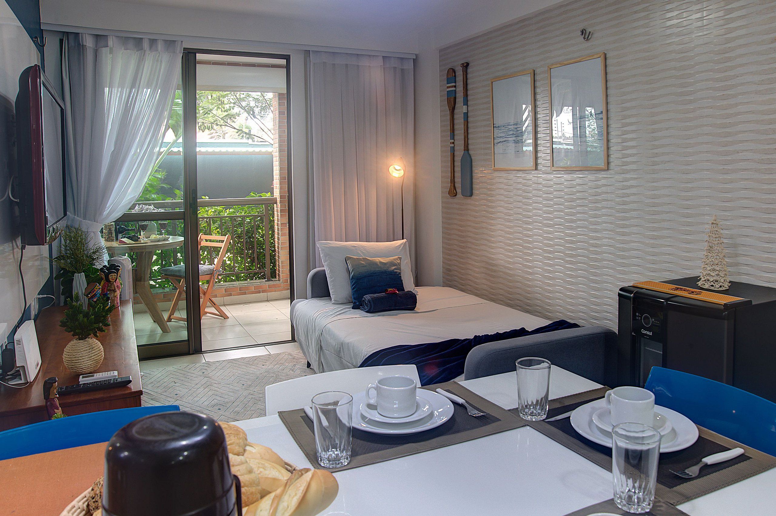 A hotel room with a bed, a desk with a computer, a dining table set for two, and a view of the outdoors.