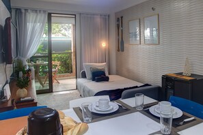 A hotel room with a bed, a desk with a computer, a dining table set for two, and a view of the outdoors.