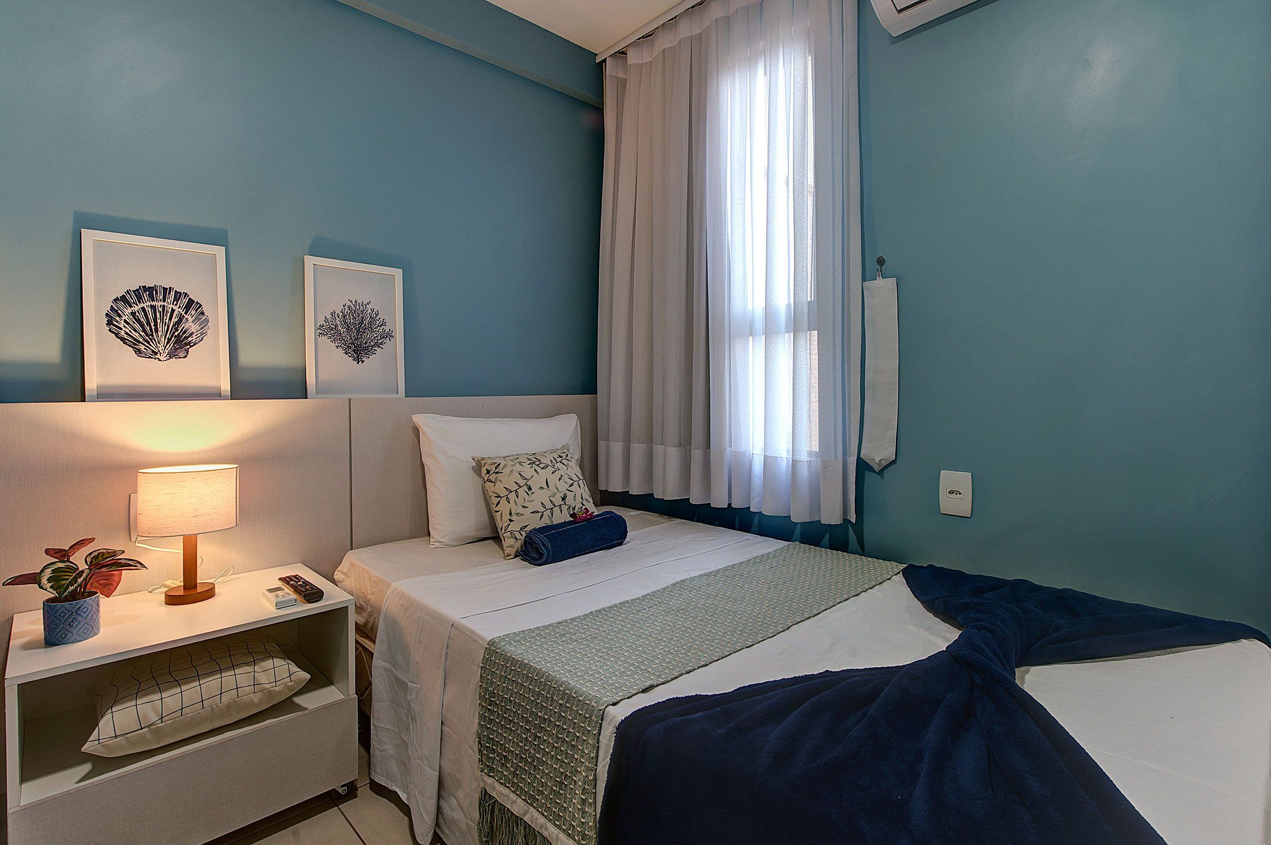 Quarto de hotel com cama, mesa de cabeceira com abajur, planta e obra de arte na parede.