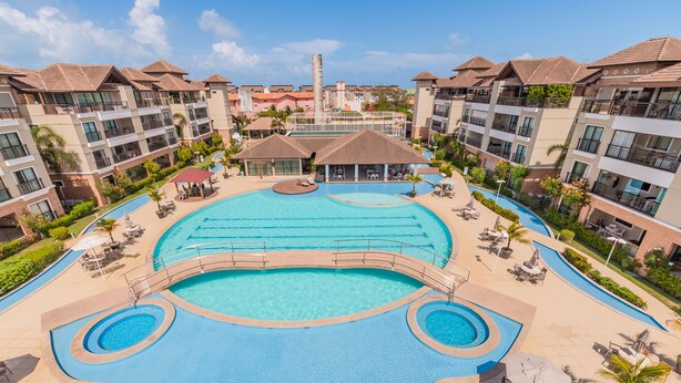 A large outdoor swimming pool surrounded by lounge chairs and umbrellas, with multiple hotel buildings in the background. - Apart em Porto das Dunas por Carpediem (Aquiraz)
