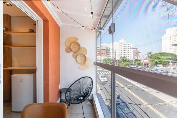 A balcony with a view of a city street, a black chair, and a refrigerator. - Coliving Vila Mariana (São Paulo)