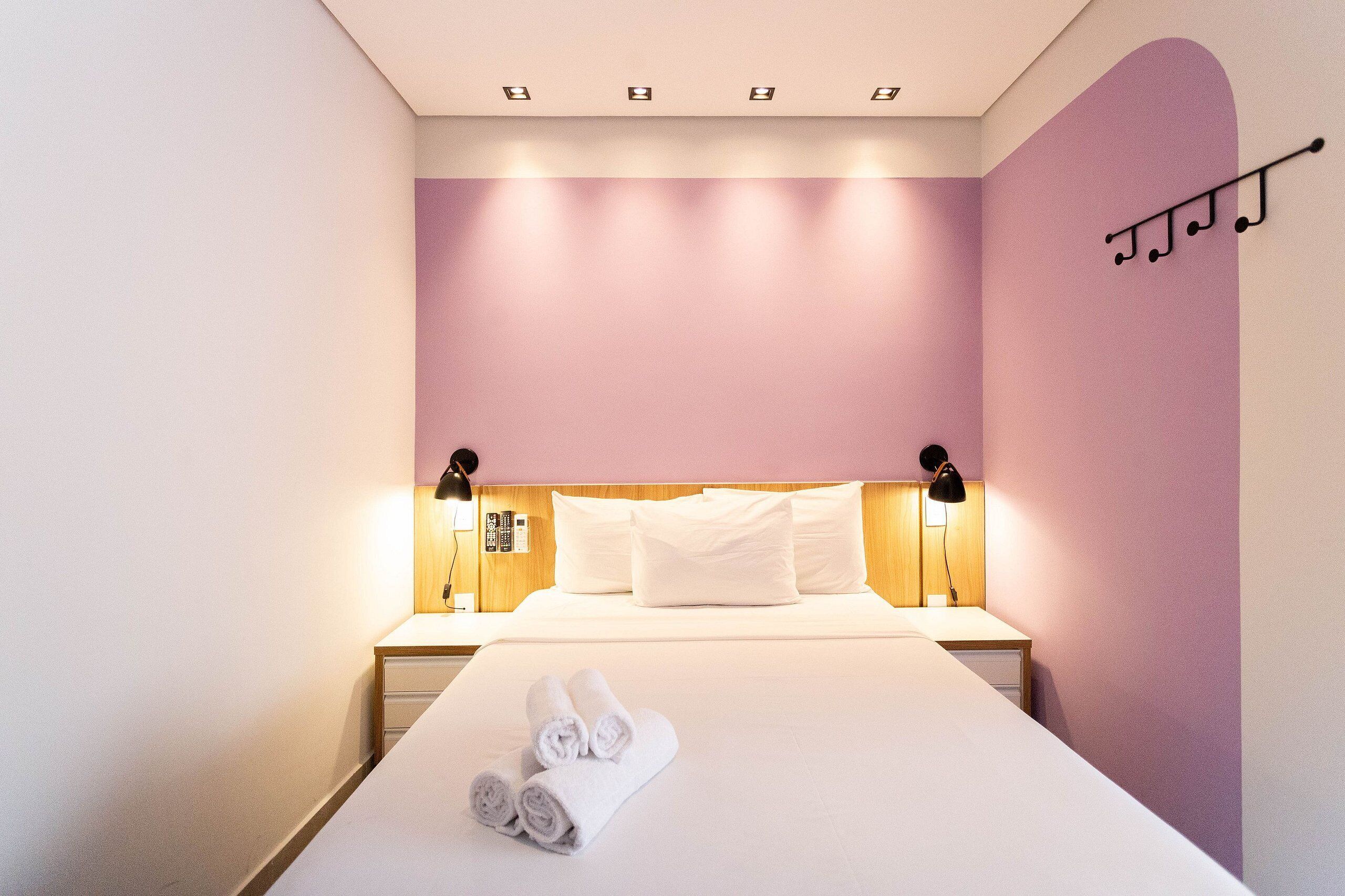 A neatly made bed with rolled towels, a wooden headboard, bedside lamps, and a wall-mounted music stand.