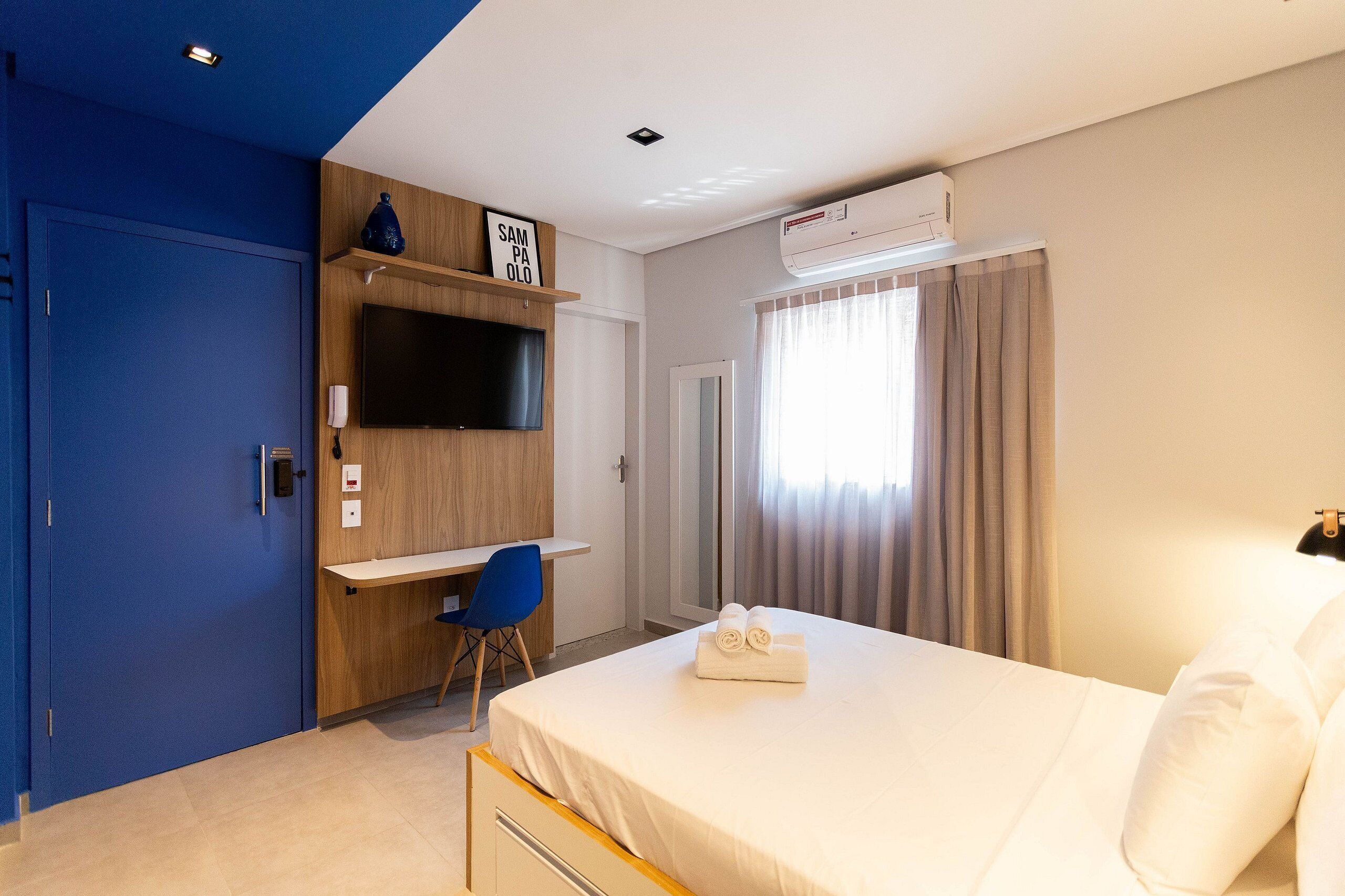 A hotel room with a bed, a desk, a TV, and an air conditioning unit.