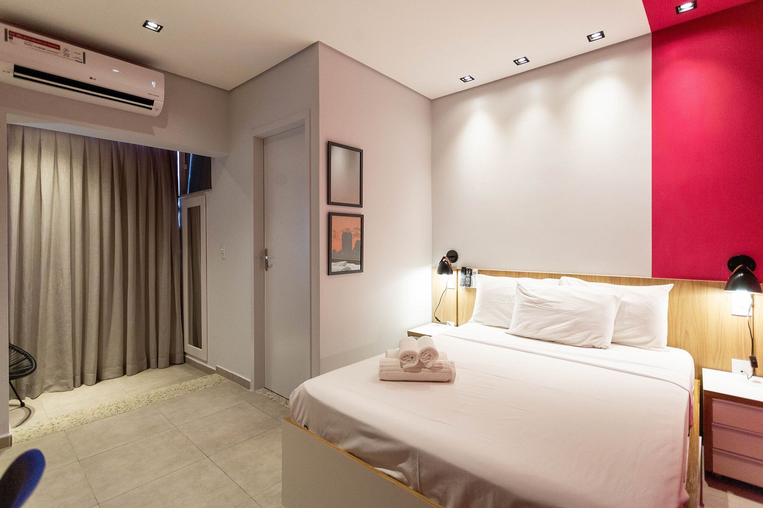 A hotel room with a bed, bedside lamps, a nightstand, a desk, an air conditioner, and a window with curtains.