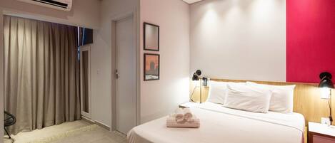 A hotel room with a bed, bedside lamps, a nightstand, a desk, an air conditioner, and a window with curtains.
