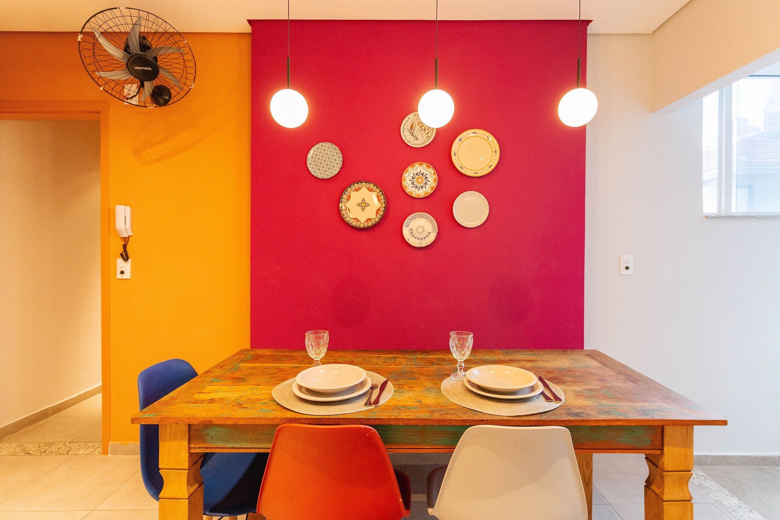 A dining area with a wooden table set for two, surrounded by colorful chairs, and a red wall with decorative plates.
