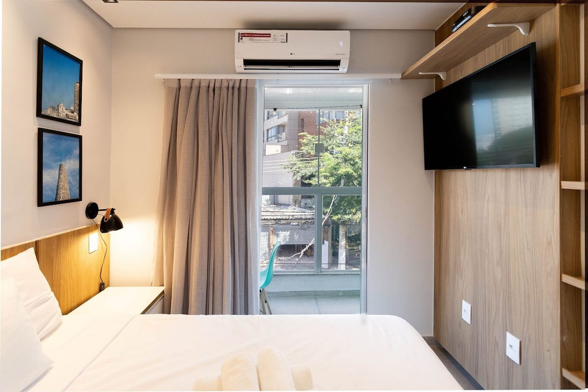 Une chambre d’hôtel avec un lit, un téléviseur fixé au mur, un appareil de climatisation et une vue sur le balcon extérieur.