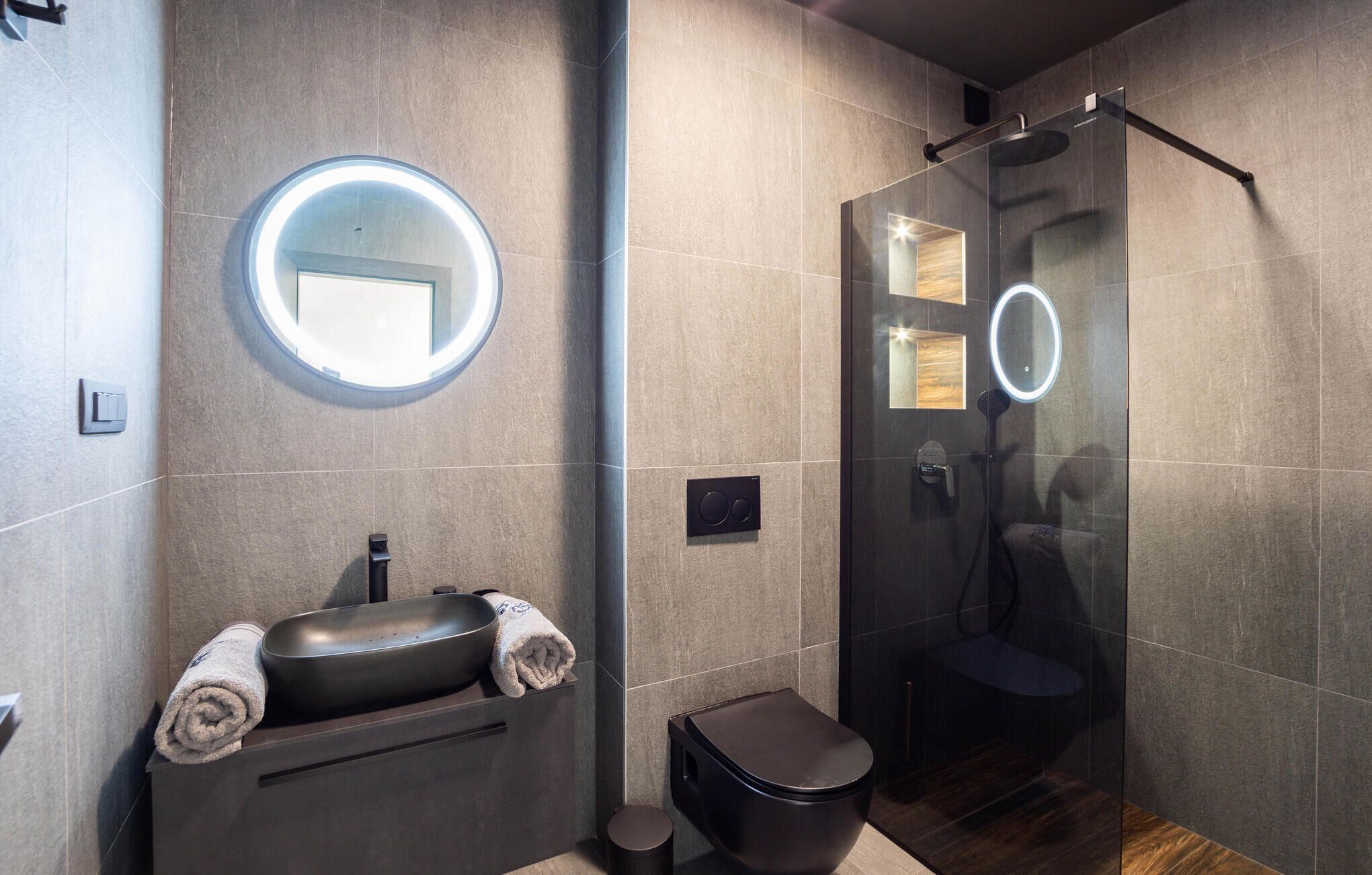 A modern bathroom with a round mirror, a sink, a toilet, and a glass-enclosed shower area.