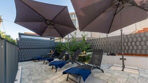 Een terras buiten met vier loungesessels en twee grote parasols, een betegelde vloer en een metalen hek op de achtergrond.