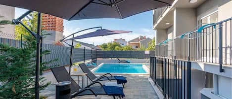 Una piscina con sillas de descanso y una zona cubierta, rodeada por una valla y edificios.