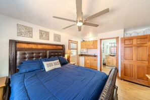 Una camera da letto con un letto grande, una ventola a soffitto, armadietti in legno e vista sulla cucina.