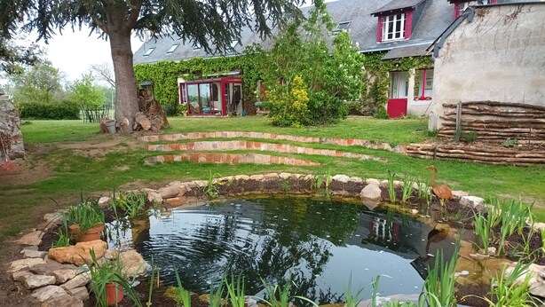 Een vijver omringd door rotsen en planten, met een huis op de achtergrond.