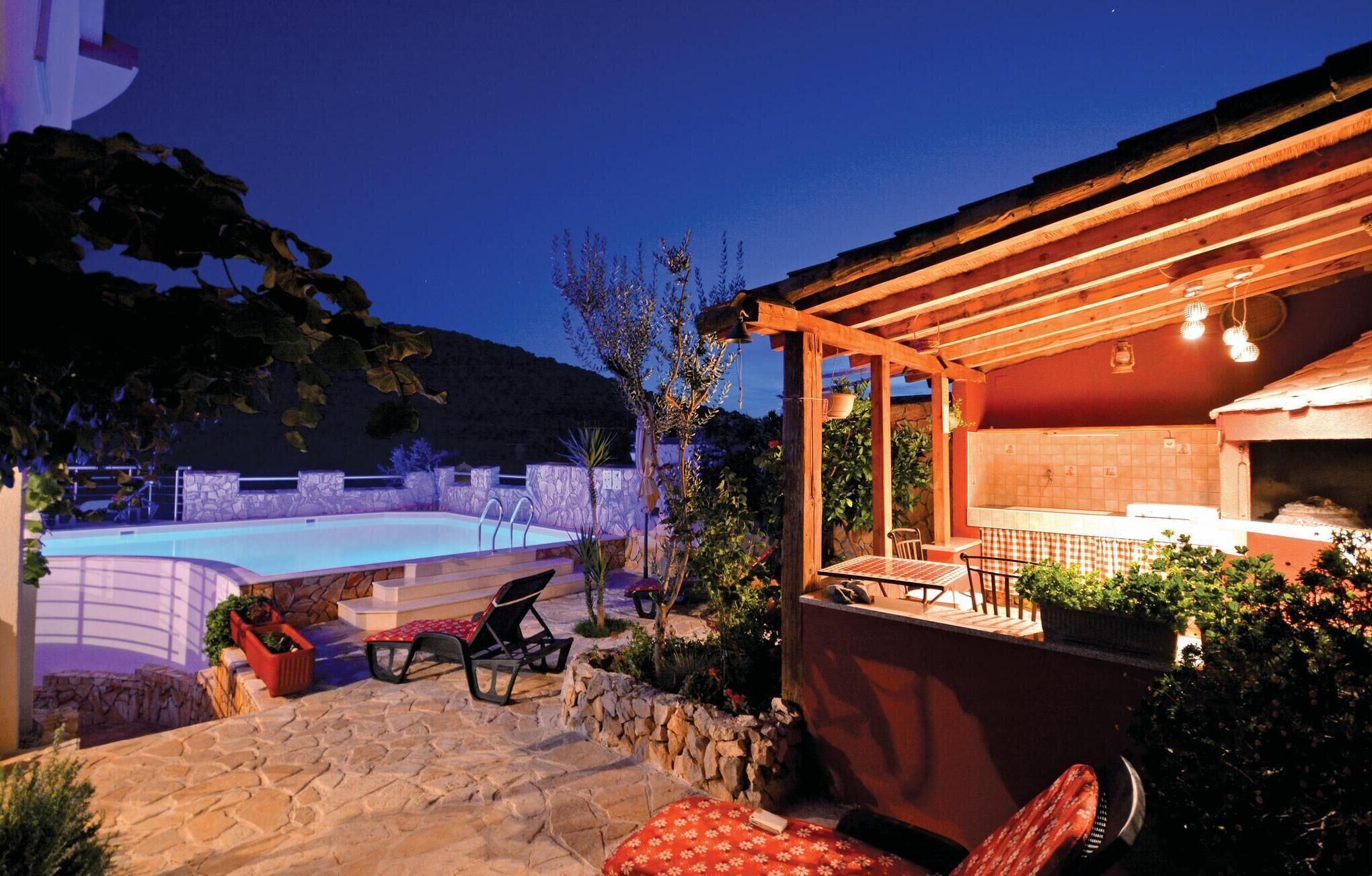 A stone pool with lounge chairs, a wooden pergola, and a kitchen area with a tiled backsplash.
