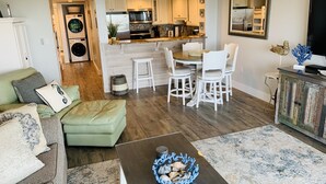 Un soggiorno con un divano verde, un tavolino da caffè con una ciotola decorativa, un'area pranzo con sedie bianche e una cucina con elettrodomestici moderni.
