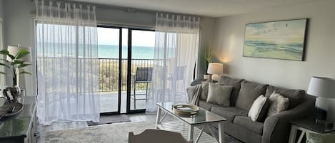 A living room with a balcony overlooking the sea, a sofa, a coffee table, and a lamp.