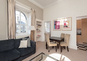 Ein Wohnzimmer mit dunkelgrauem Sofa, einem Glastisch, einem Esstisch mit Stühlen und Blick auf die Stadt durch große Fenster.