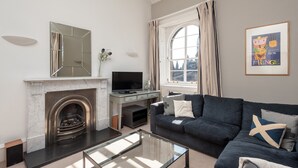 A living room with a fireplace, a television, a sofa, and a large window. - Fantastic corner 2 bedroom property with astounding views of the famous historic Royal Mile. (Edinburgh)