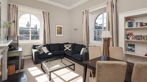 A living room with a dark blue sofa, a glass coffee table, a wooden side table, and a bookshelf filled with books. - Fantastic corner 2 bedroom property with astounding views of the famous historic Royal Mile. (Edinburgh)