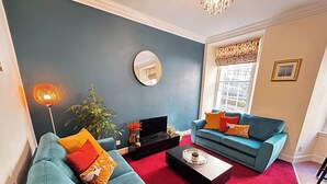 A living room with teal sofas, a red carpet, a black entertainment unit, and a large window with patterned curtains. - Economical 2 bedroom property just off the famous Royal Mile (Edinburgh)