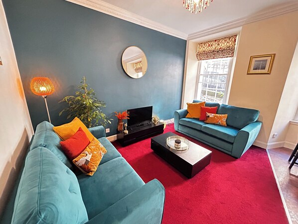 A living room with teal sofas, a red carpet, a black entertainment unit, and a large window with patterned curtains. - Economical 2 bedroom property just off the famous Royal Mile (Edinburgh)