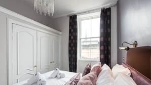 A neatly made bed with white linens and pink pillows, a chandelier, a window with curtains, and a door with a handle. - Incredible 1 bedroom + sofabed on the famous Royal Mile 200 metres from Edinburgh Castle (Edinburgh)