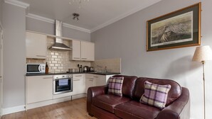 A modern kitchen with white cabinets, a microwave, and an oven. There is a brown leather sofa with plaid cushions in the living area.