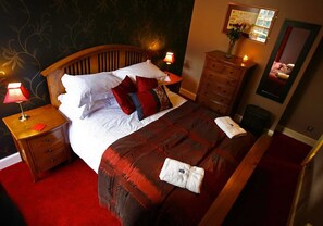 A neatly made bed with a red and white duvet, two red pillows, and a wooden headboard. There are two bedside tables with lamps, a dresser, and a mirror on the wall.