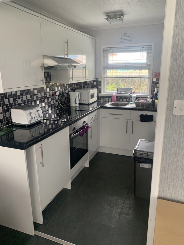 Compact kitchen with white cabinetry, black and white tiles, microwave, toaster, oven, and window above the sink.