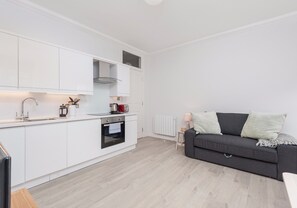 A modern kitchen with white cabinetry, a built-in oven, and a grey sofa in the living area.