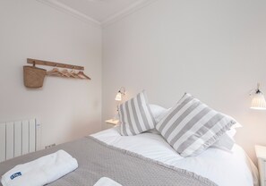 A neatly made bed with a grey blanket, white pillows, and a white towel on the side. There is a wooden coat rack with a woven bag and wooden hangers. Two wall-mounted lamps provide lighting.