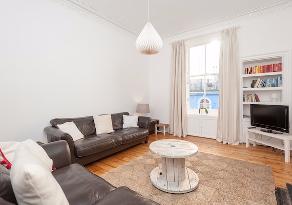 A living room with a brown leather sofa, a round coffee table, a bookshelf, a television, and a window with curtains.