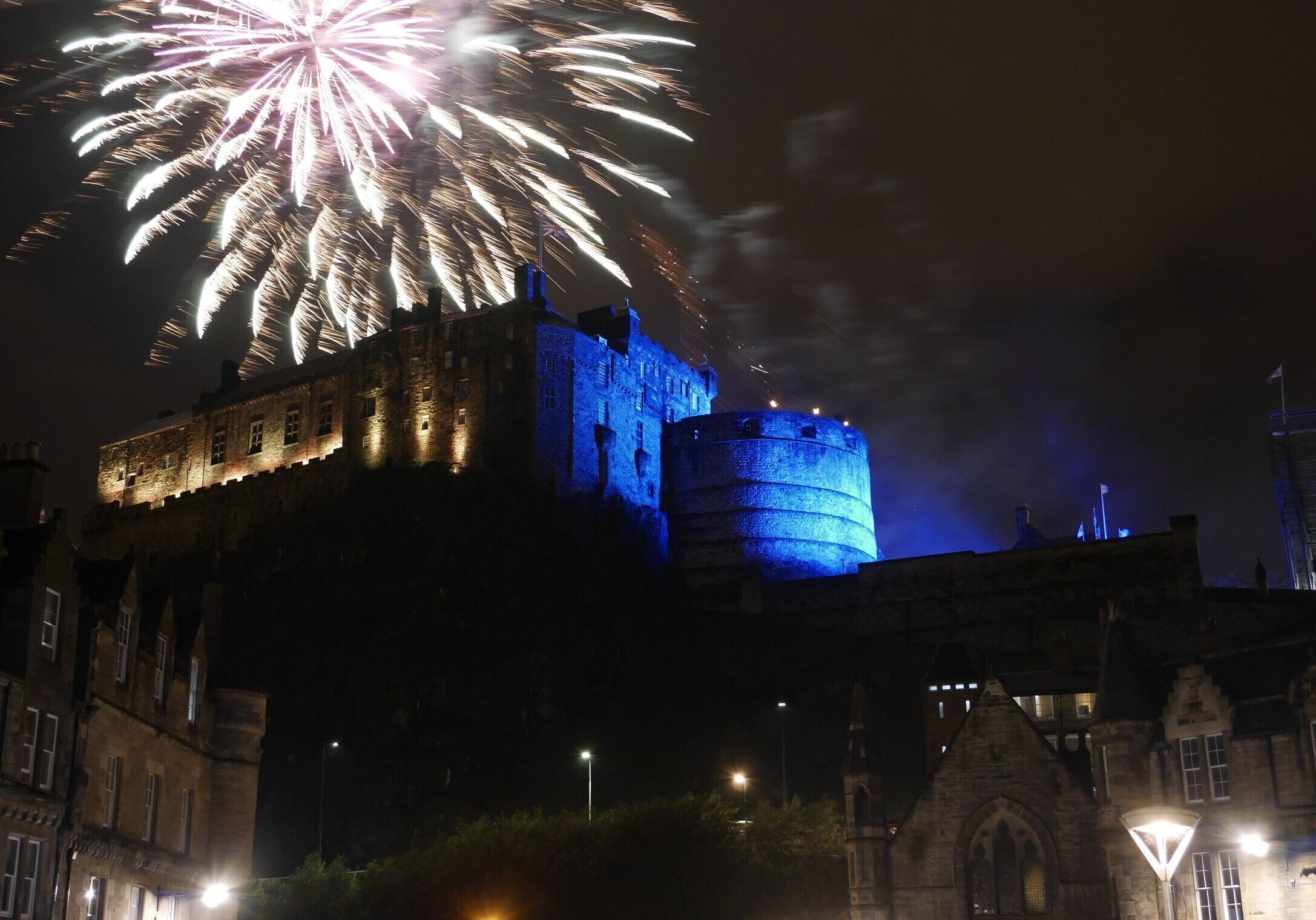 A historic castle with a round tower, illuminated by blue lights, and fireworks in the background.
