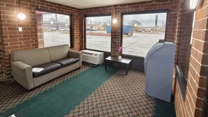 A room with a sofa, a small table with a vase, a heater, and a large window with a view of a parking lot.