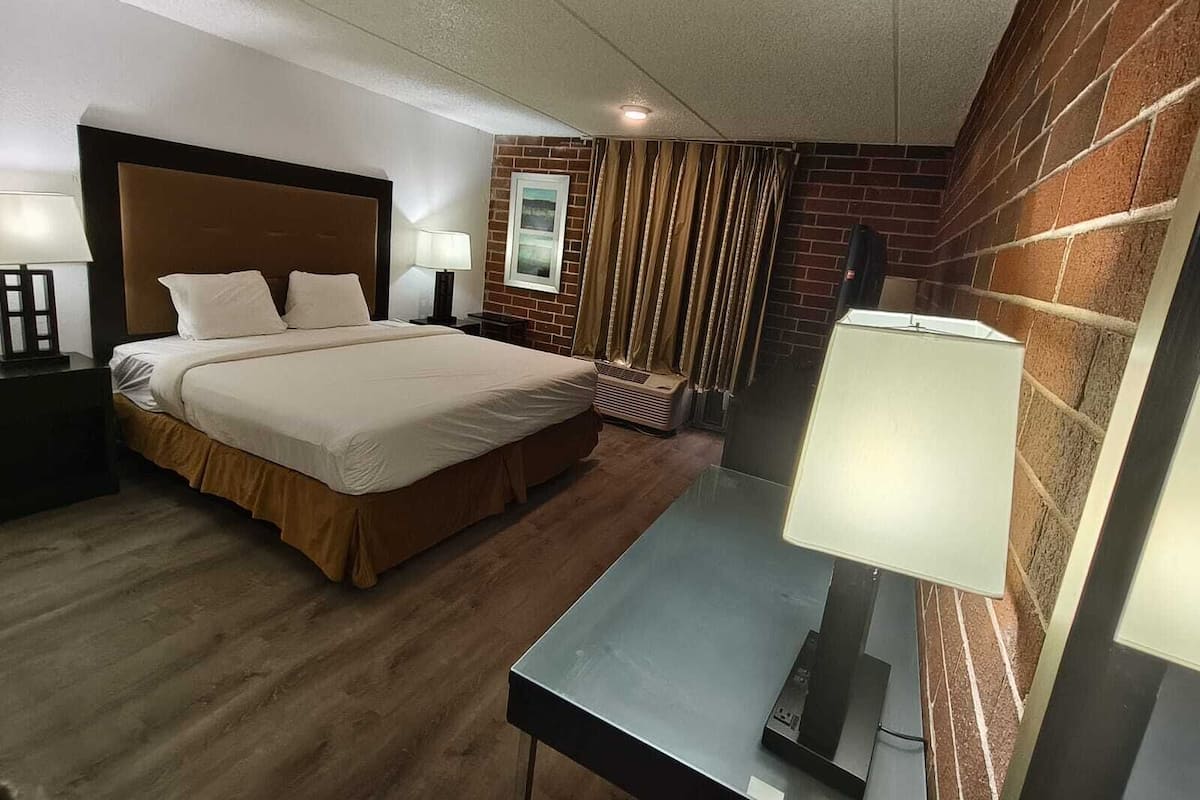 Una habitación de hotel con cama, mesitas de noche, escritorio, lámpara, ventana con cortinas y pared de ladrillo.