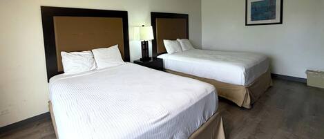Une chambre d’hôtel avec deux lits simples, une table de chevet avec une lampe, un tableau encadré au mur et un plancher en bois.