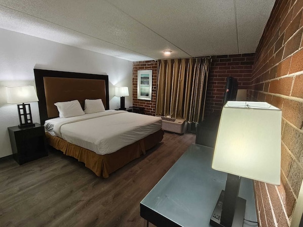 A hotel room with a bed, two bedside tables with lamps, a window with curtains, a brick wall, and a small heater. - Plaza Hotel By OYO Calumet Park Near Alsip I 57 (Calumet Park)