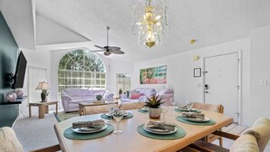 Un coin repas avec une longue table dressée pour quatre personnes, entourée de chaises rembourrées. Un ventilateur de plafond et un lustre sont visibles au-dessus de la table.