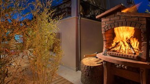 Chimenea de ladrillo al aire libre con fuego encendido, rodeada por una terraza de madera y una planta de bambú.