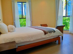 Ein Schlafzimmer mit einem großen Bett, zwei Stühlen und Blick auf Grün hinter den Fenstern.