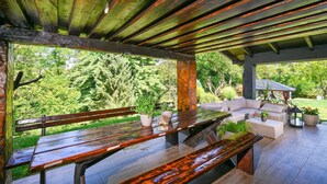 Une terrasse extérieure avec des bancs en bois et un coin salon confortable, le tout entouré de verdure.