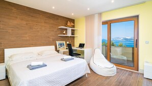 A modern bedroom with a large bed, a desk, and a chair. There is a view of the sea through a sliding glass door.