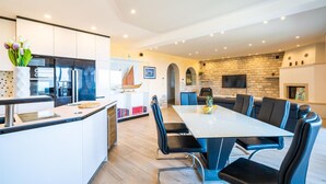 A modern kitchen with a central island, a dining area with a long table and chairs, and a living area with a fireplace and a TV.