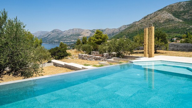 A swimming pool with clear blue water, surrounded by a stone wall and a view of mountains and a lake.