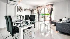 Un salon moderne avec une table à manger en verre, des chaises noires, un canapé gris et une vue sur l’extérieur.