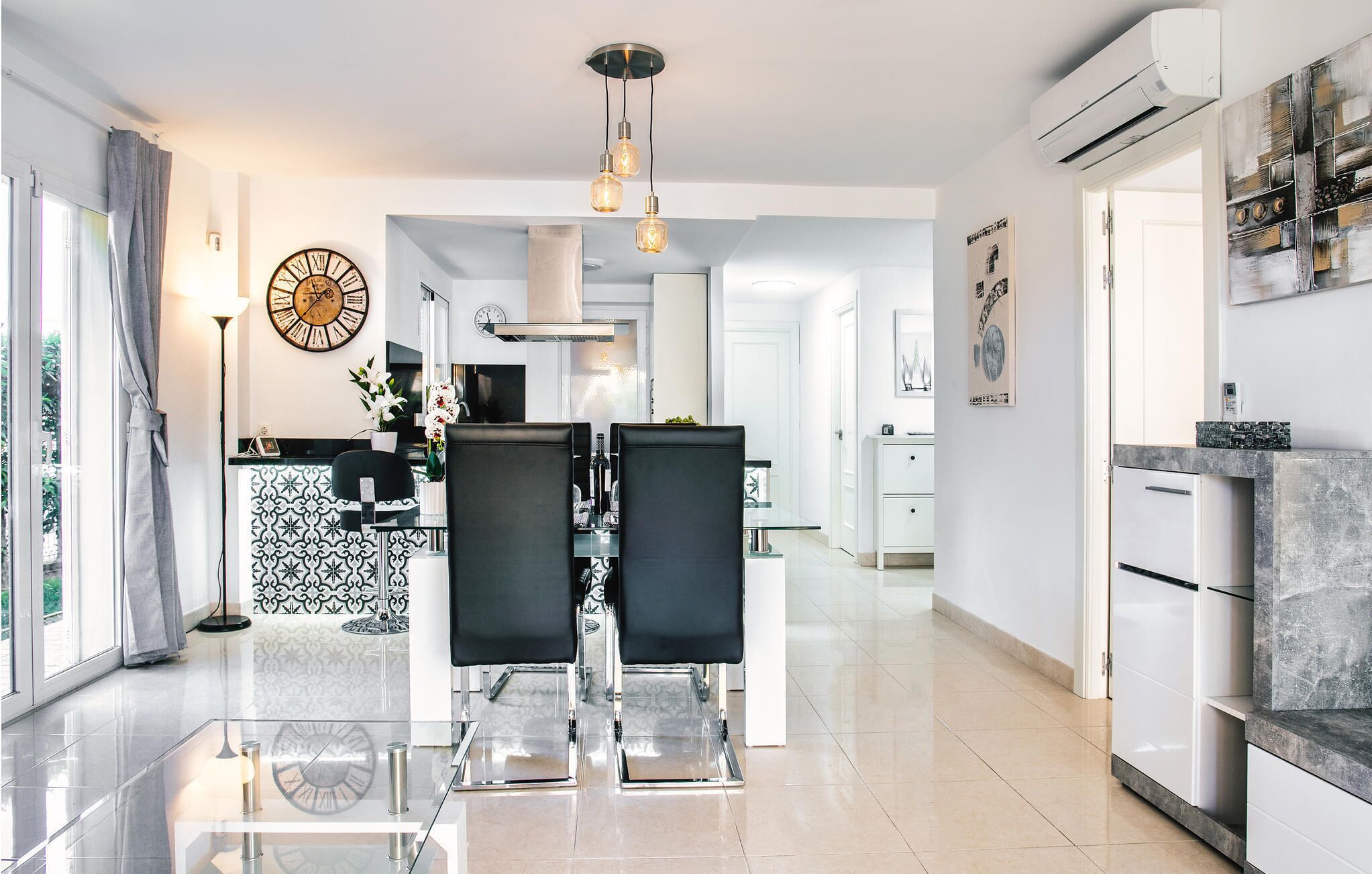 Une cuisine moderne avec un coin repas, agrémentée d’une grande horloge, de luminaires suspendus et offrant une vue sur l’extérieur.