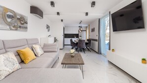 Moderni olohuone, jossa on harmaa sohva, sohvapöytä ja ruokailutila, jonka yllä on koristeellinen kattokruunu.